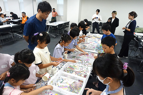長いテーブルを前に作業をしている子供たちとそれを見守っている親たち